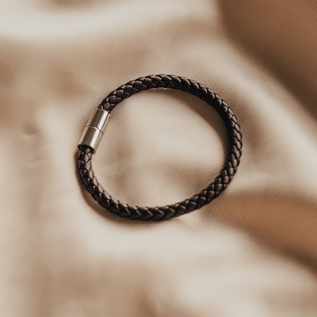 Mørkebrunt flettet lærarmbånd med glatt, rund magnetlås i sølvfarget metall, vist mot hvit bakgrunn.