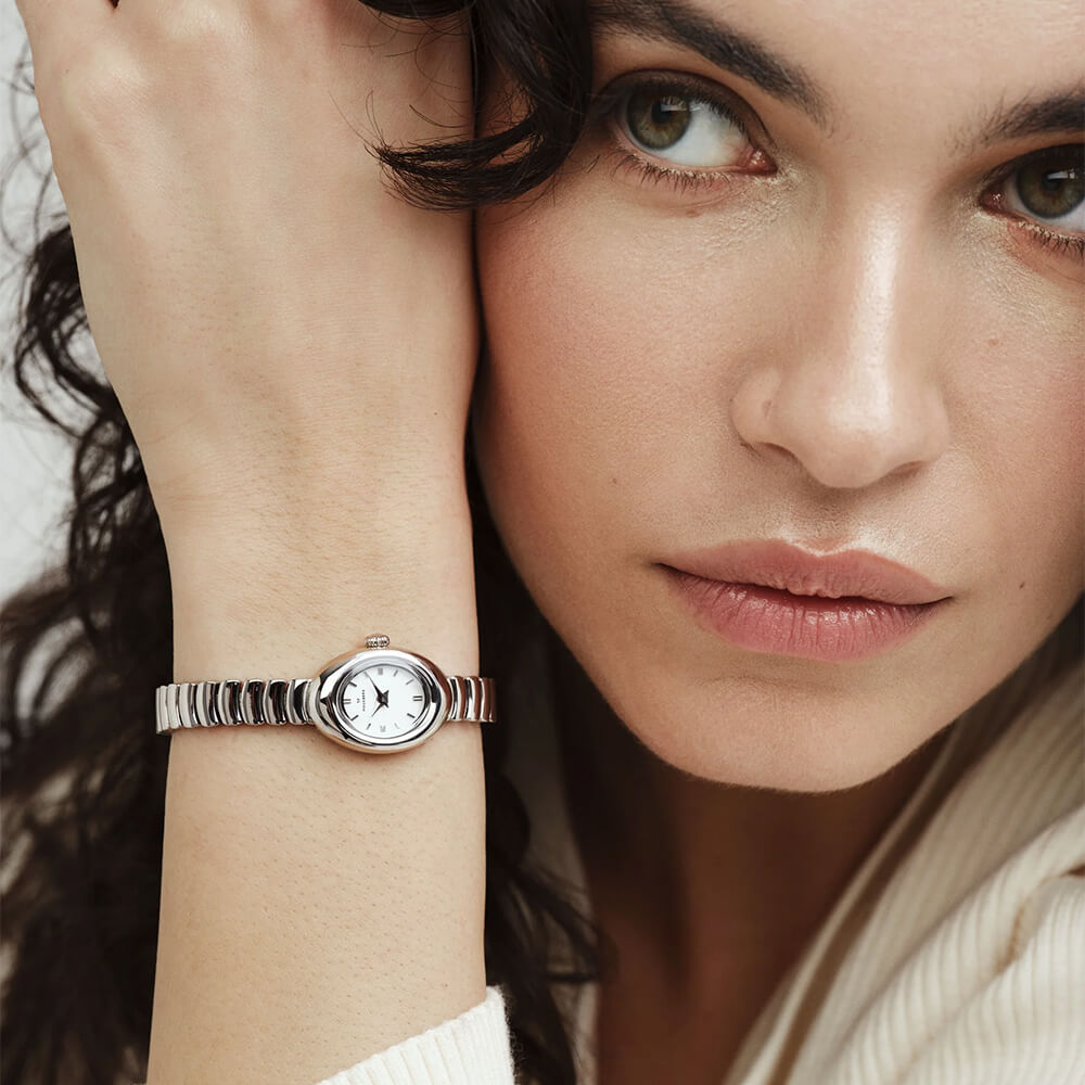 Sølvfarget armbåndsur med oval urkasse, hvit urskive og elastisk lenke i metall, vist mot hvit bakgrunn med svak skygge under.