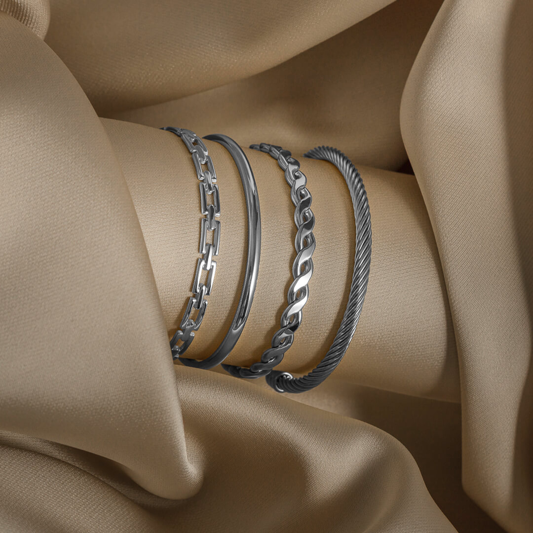 Åpen armring med lenkeinspirert mønster i sølvfarget metall, plassert på hvit bakgrunn med svak skygge under.