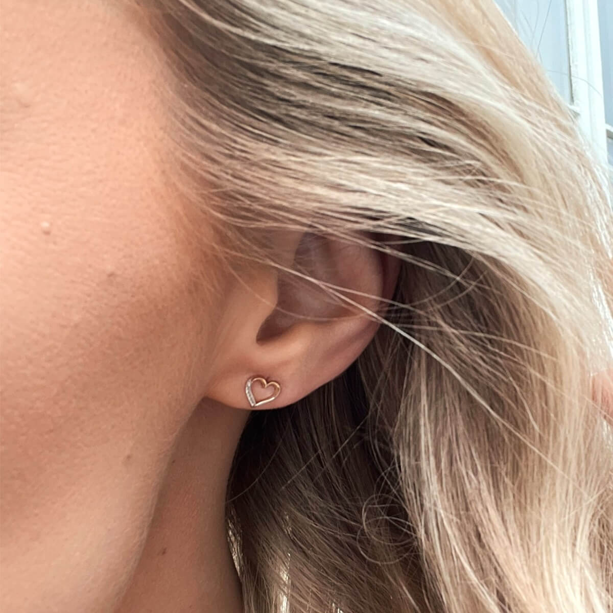 Et par åpne hjerteformede øredobber i gullfarget metall med en rekke små, klare steiner langs den ytre kanten.