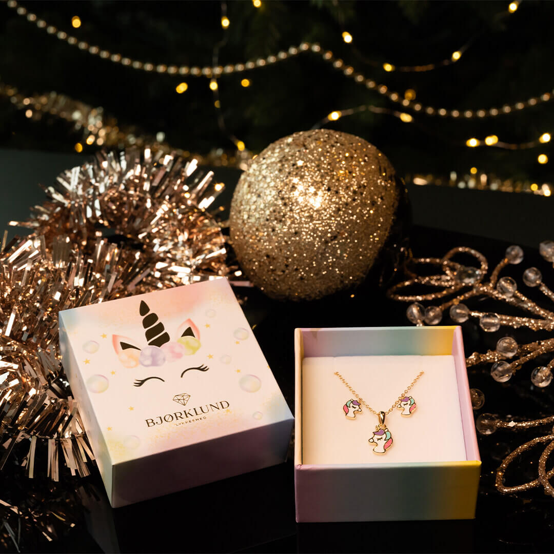 Et par små øredobber formet som enhjørninger i gullfarge med fargerik man og horn, presentert i en hvit gaveeske med regnbuefarget kant. 