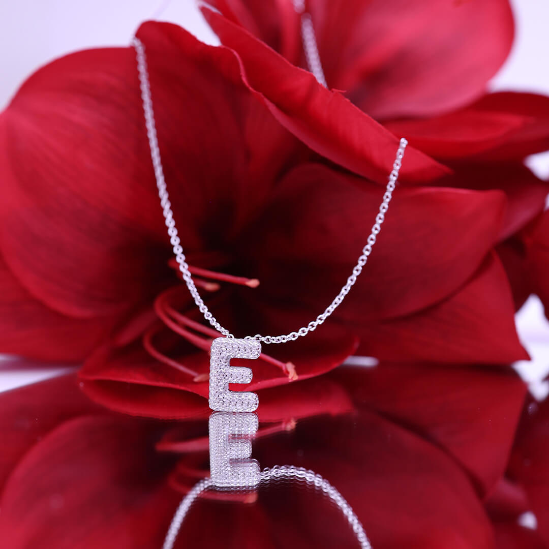 Anheng i form av bokstaven E, pavébesatt med små stener, hengt på et sølvkjede, avbildet mot hvit bakgrunn.