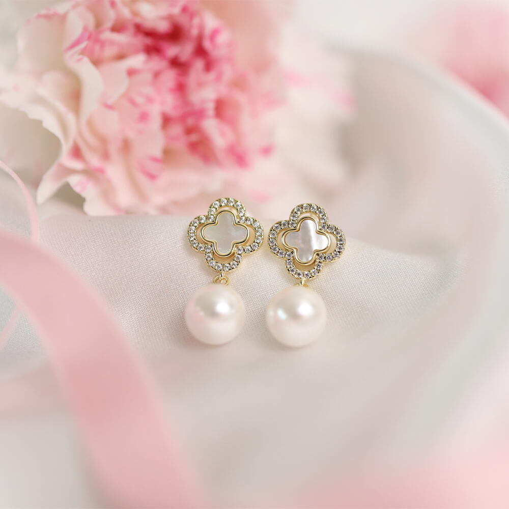 Et par gullfargede øredobber med kløverformede detaljer fylt med hvit emalje og små steiner rundt kanten, samt store hvite perler som henger under, avbildet mot hvit bakgrunn.