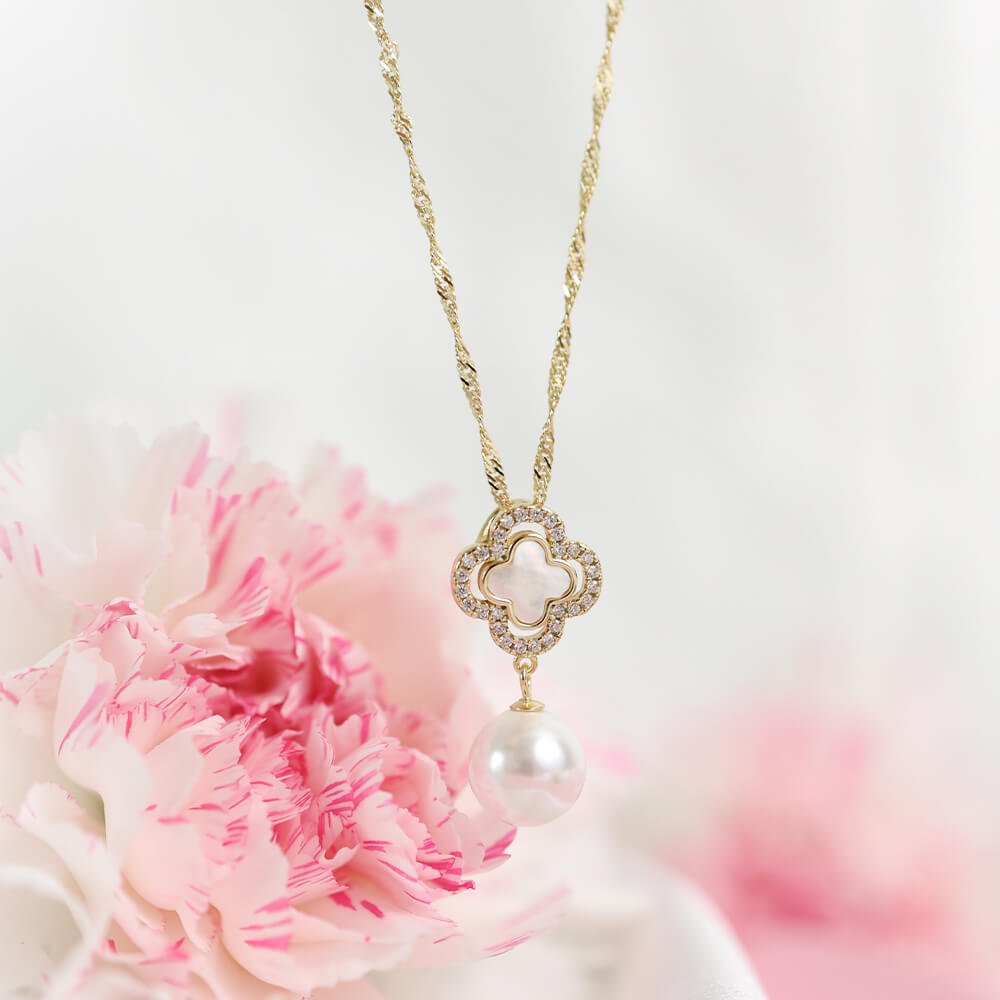 Et gullkjede med et dekorativt anheng som har kløver med små steiner og en stor, rund perle hengende nederst, avbildet mot hvit bakgrunn.