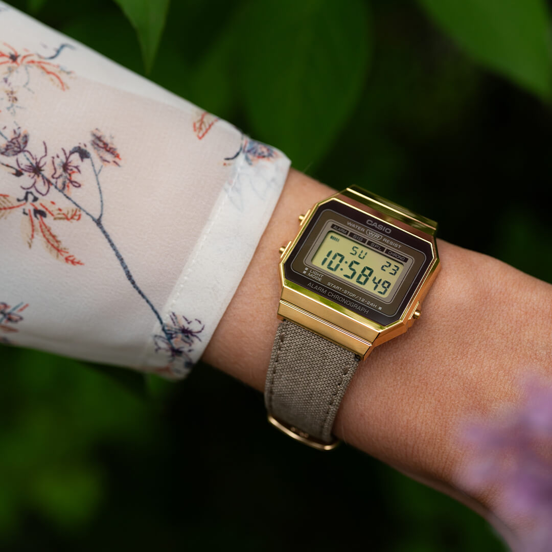 Digitalklokke med brun tekstilrem og gullfarget urkasse i rektangelform.