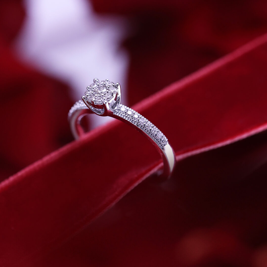 Ring med rund klynge av små diamanter i klørfatning og diamantbesatte skuldre, avbildet mot hvit bakgrunn.