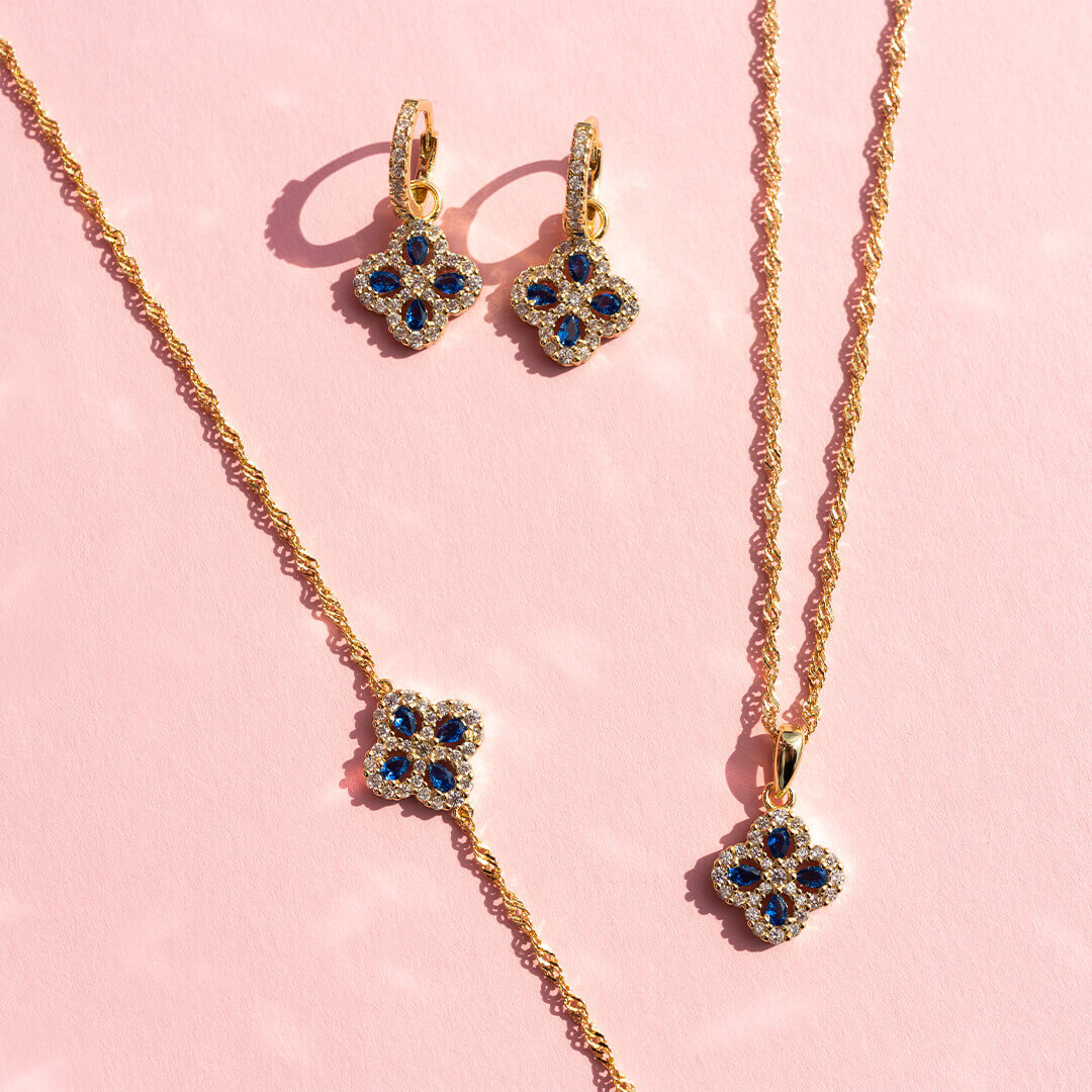 Et par forgylte øredobber med hengende kløverformet anheng dekorert med blå og klare stener, samt små klare stener langs ørekroken.