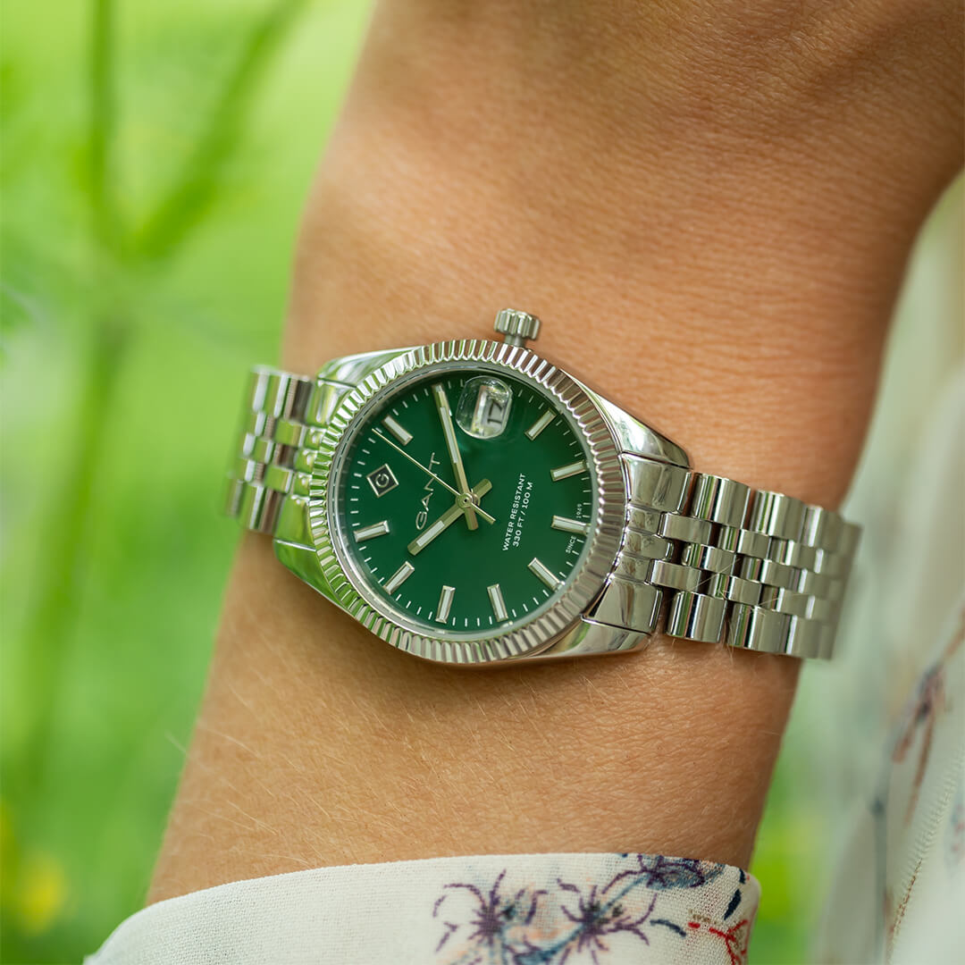 Sølvfarget klokke med sølvfargede indekser og visere og datovisning med forstørrelsesglass på en grønn urskive.