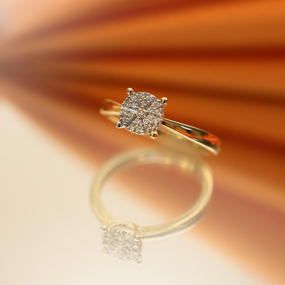 Ring i blankt gult metall med bredt bånd og sirkelrund topp dekorert med flere små, klare stener i stjernemønstret fatning.