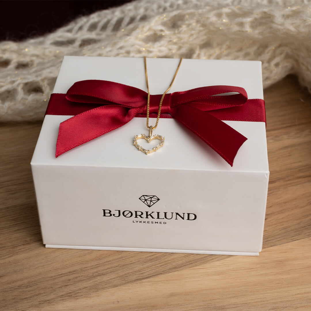 En dame med lyst hår som har på seg rød bluse, bærer på et nydelig halssmykke i gult gull med hjerteanheng.