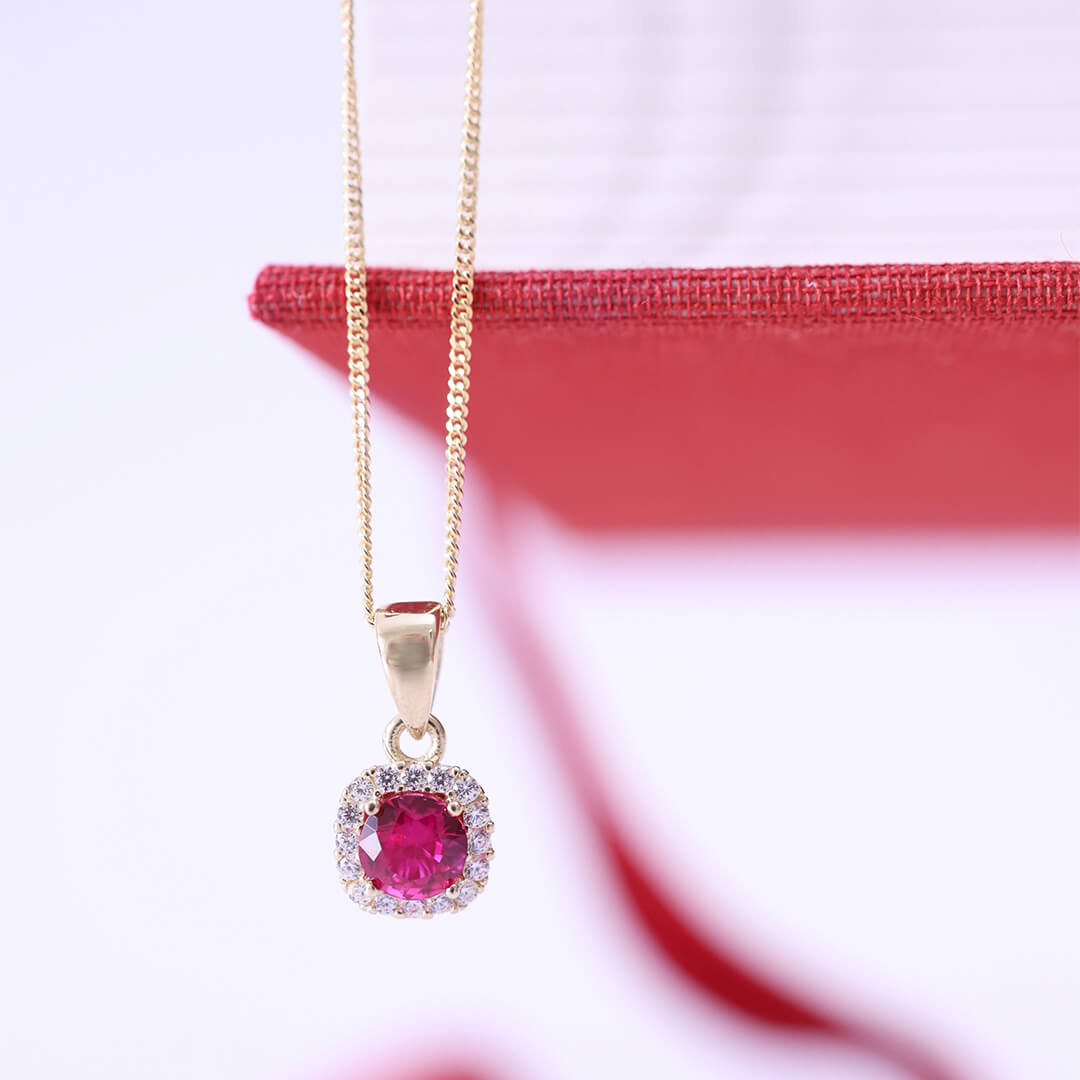 Halssmykke i gult gull med rund, rød midtsten omgitt av små hvite steiner, festet til en tynn kjede mot hvit bakgrunn.
