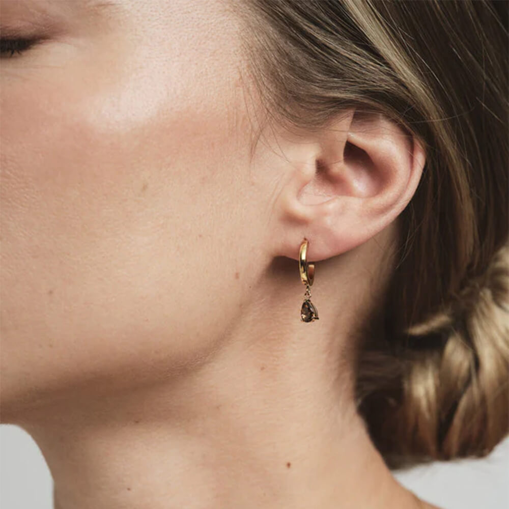 Gullfargede øreringer med dråpeformet brunrød stein som henger fritt, vist på hvit bakgrunn for produktvisning. 