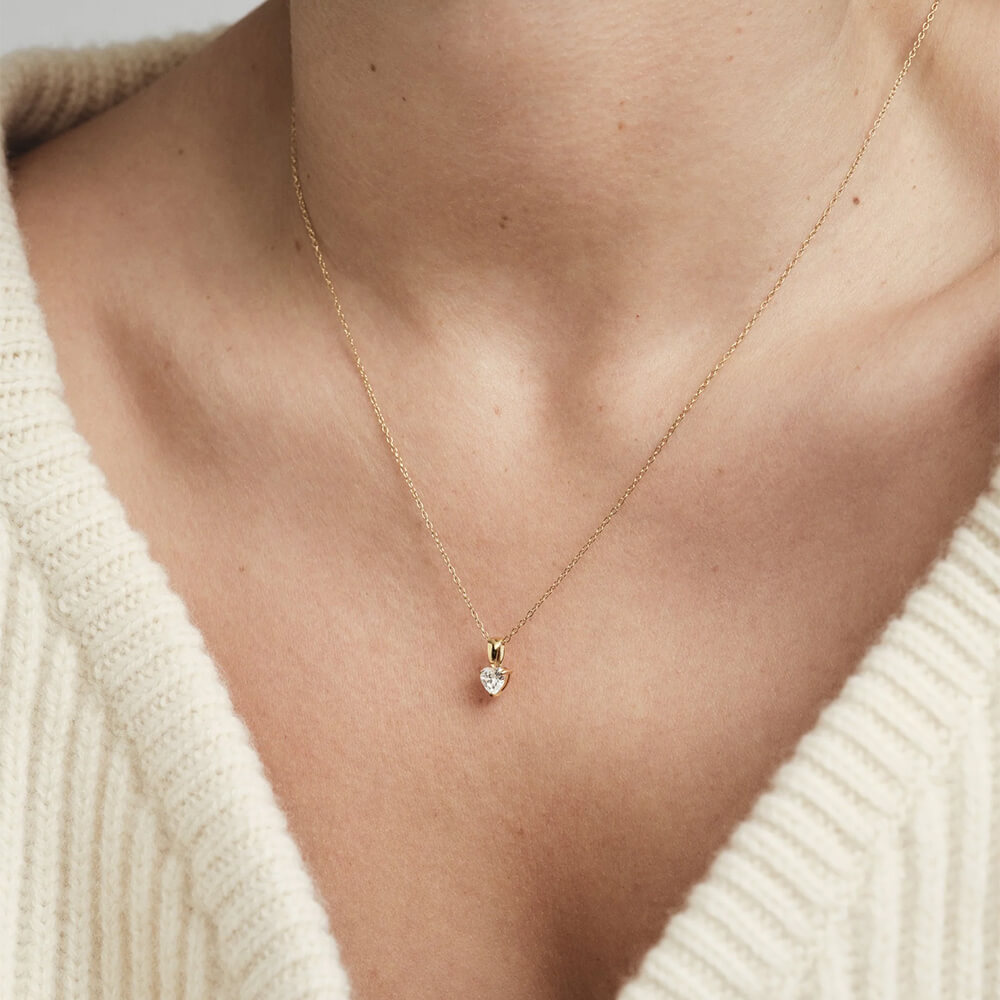 Et halskjede i gull med en tynn lenke og et hjerteformet anheng med klar stein, fotografert mot hvit bakgrunn.