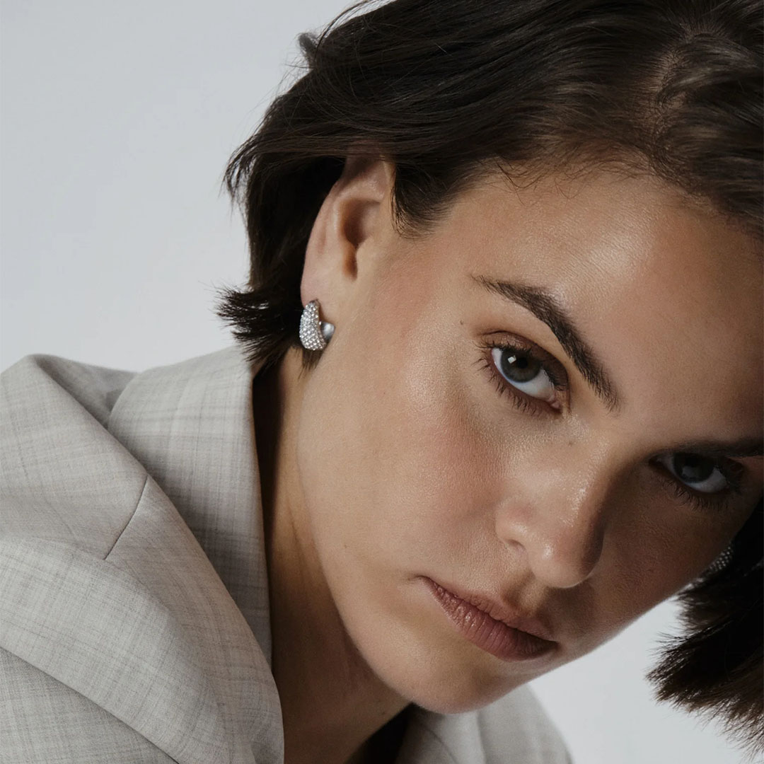 Et par øreringer i sølvfarge dekorert med hvite stener på en side.