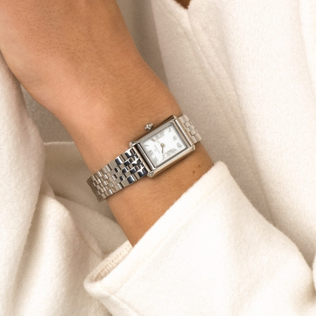 Sølvfarget dameklokke med rektangulær kasse, hvit urskive med romerske tall og treledd stållenke mot hvit bakgrunn med skygge