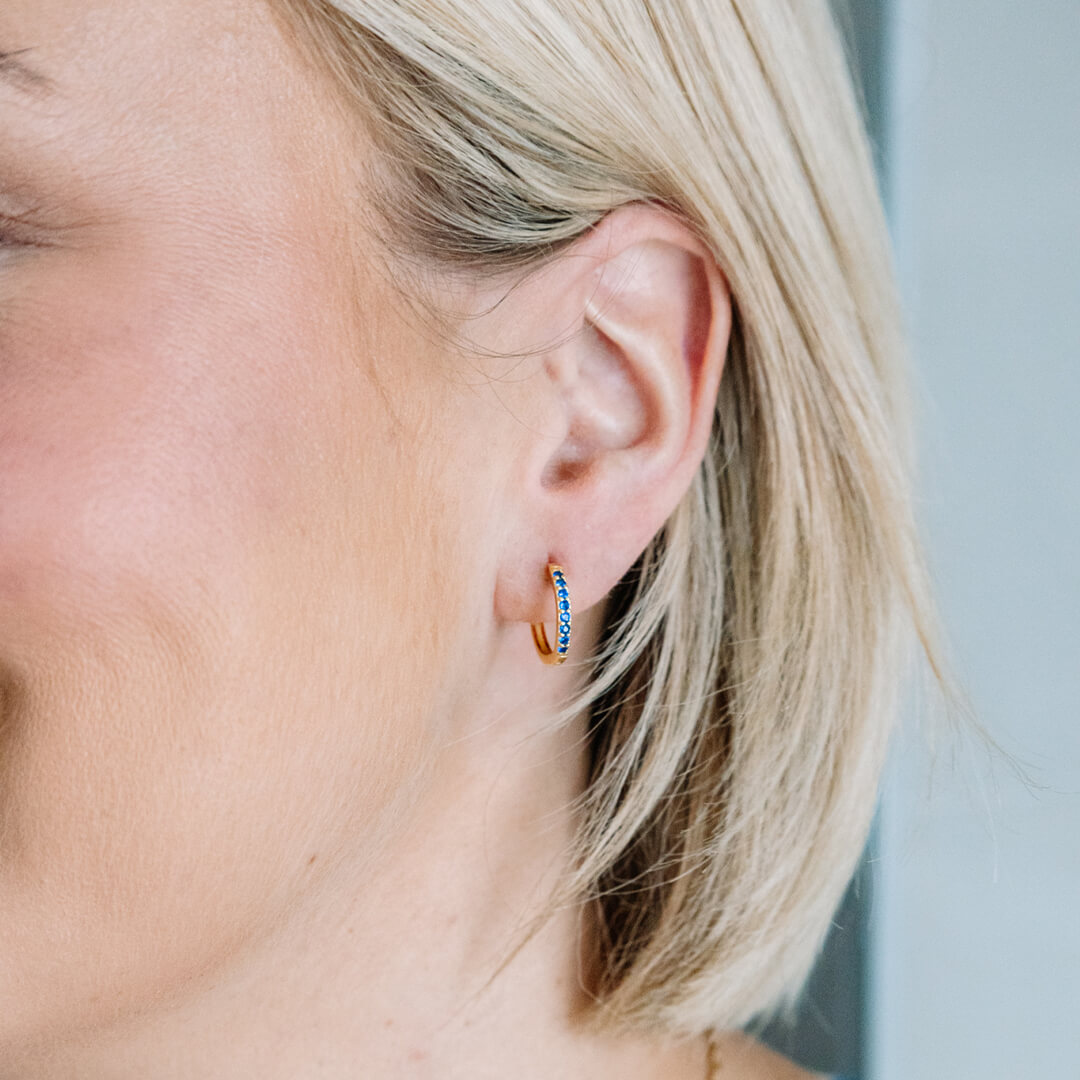 Forgylte øreringer med blå stener langs fremsiden, avbildet mot hvit bakgrunn.