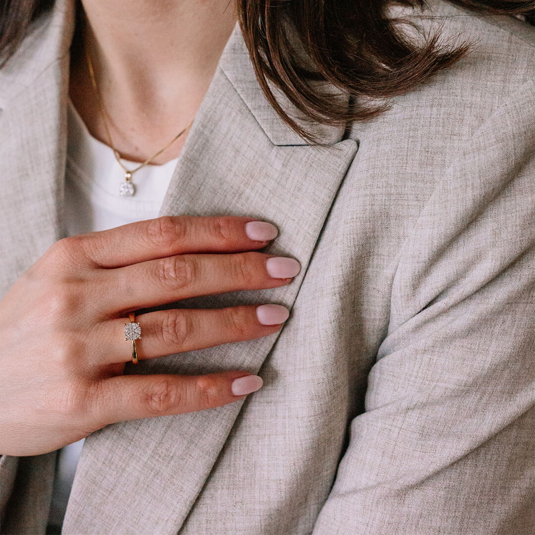 Ring i blankt gult metall med bredt bånd og sirkelrund topp dekorert med flere små, klare stener i stjernemønstret fatning.