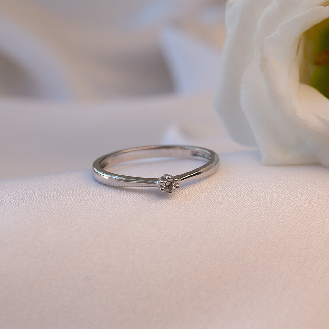 En smal ring med en liten rund diamant festet i fire klør, fotografert mot hvit bakgrunn med svak skygge under ringen.