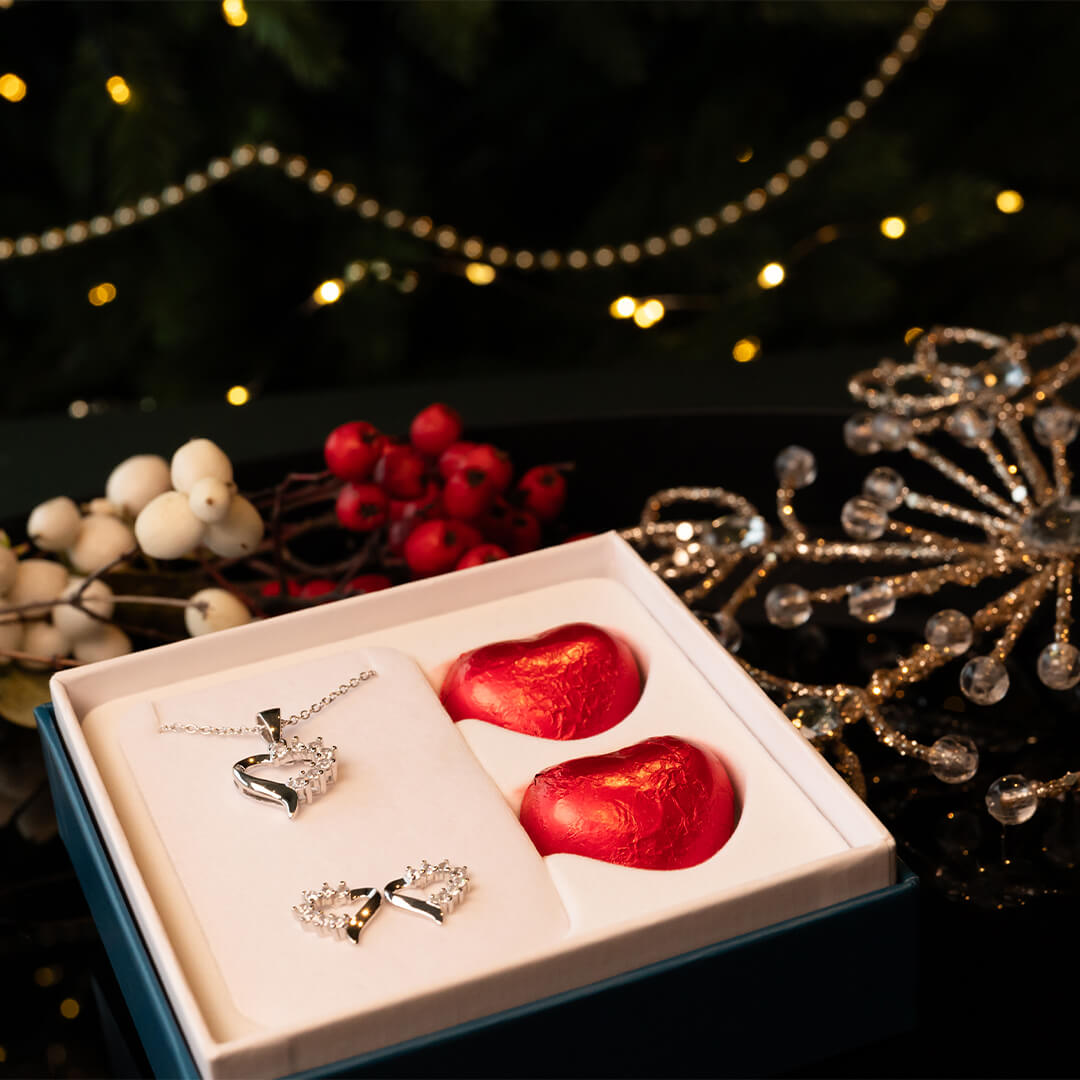 Sølvfarget smykkesett med halssmykke og creol-øredobber i asymmetrisk hjerte-design dekorert med gjennomsiktige steiner mot hvit bakgrunn