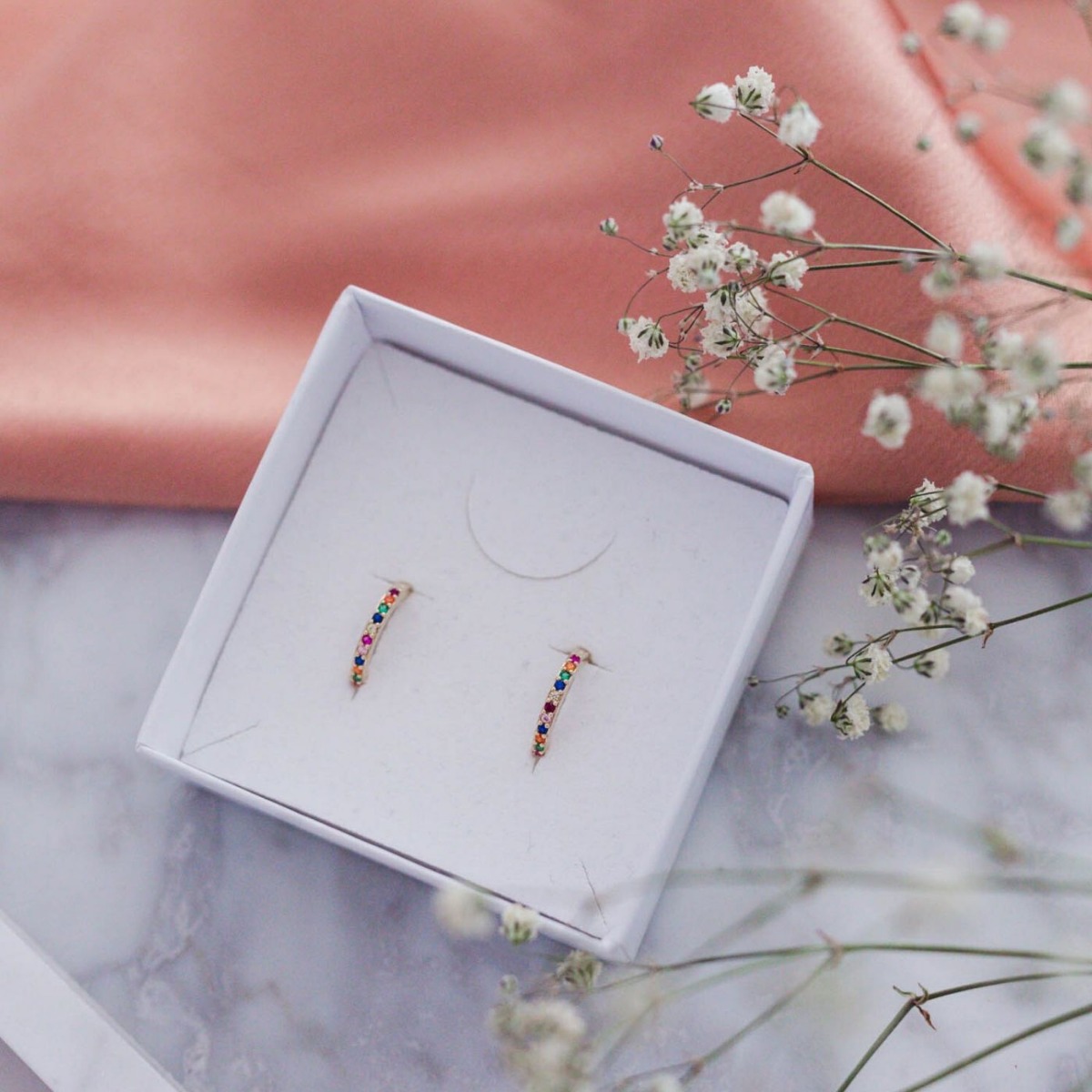 Par små gullfargede øreringer i ringform med innfelte steiner i ulike farger, vist mot hvit bakgrunn.