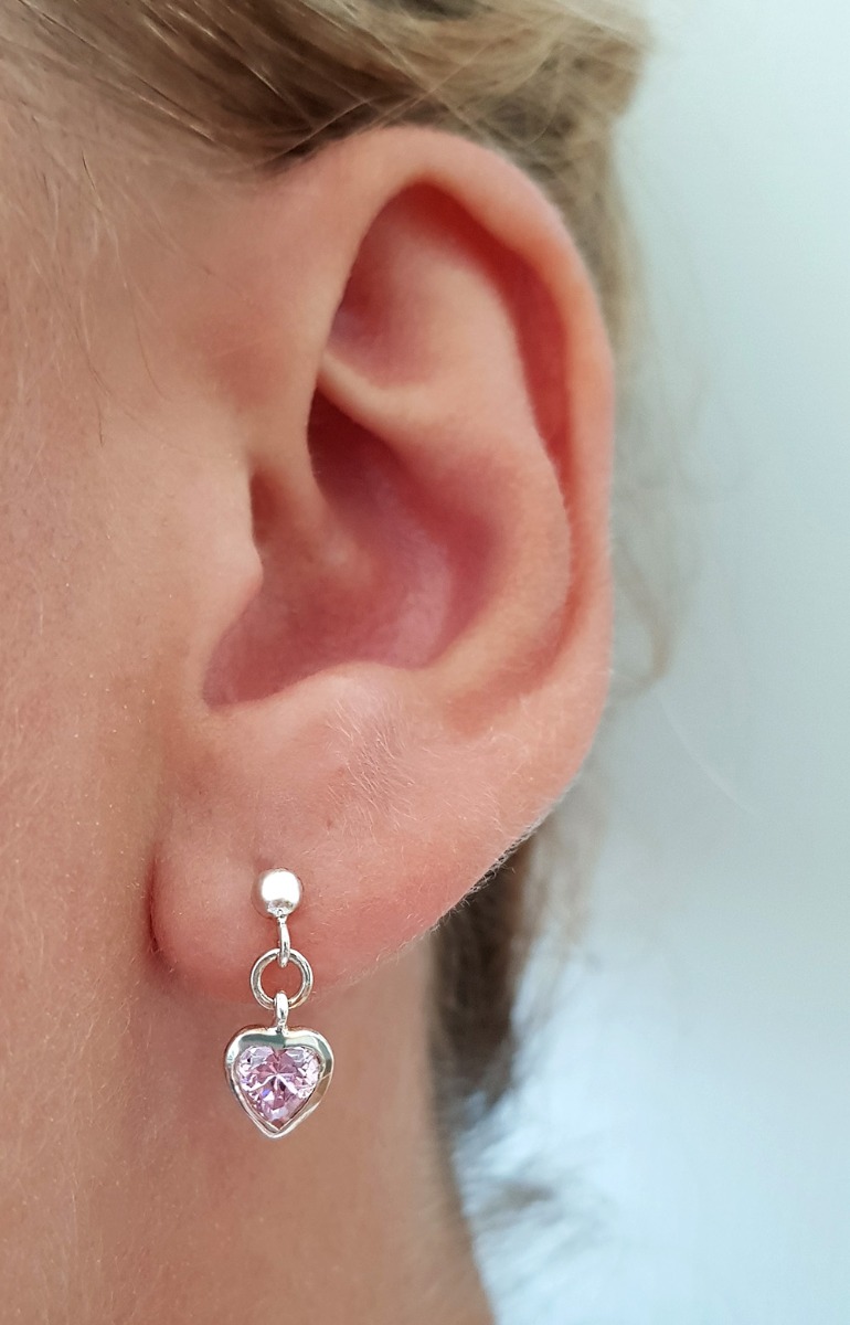 Sølvfargede øredobber med hengende hjerteformede anheng og rosa steiner, avbildet mot hvit bakgrunn.
