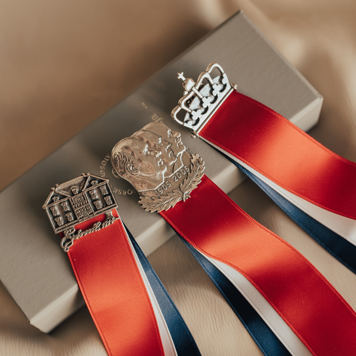 Sølvsløyfe med motiv av tre profiler og hengende bånd i rødt, hvitt og blått som symboliserer Norge.