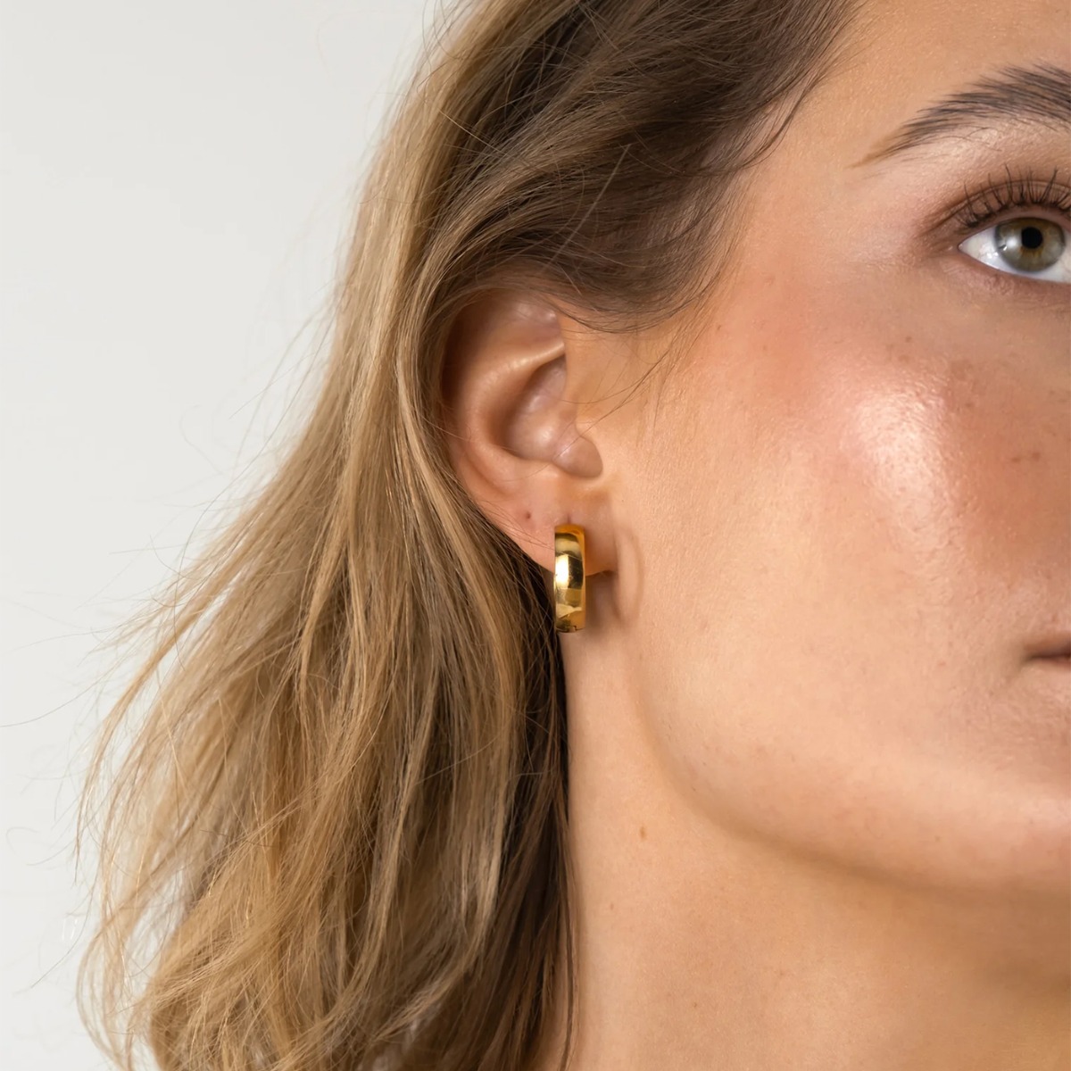 Et par små, glatte øreringer i gullfarget metall, avbildet stående oppreist mot en hvit bakgrunn med svak refleksjon under.