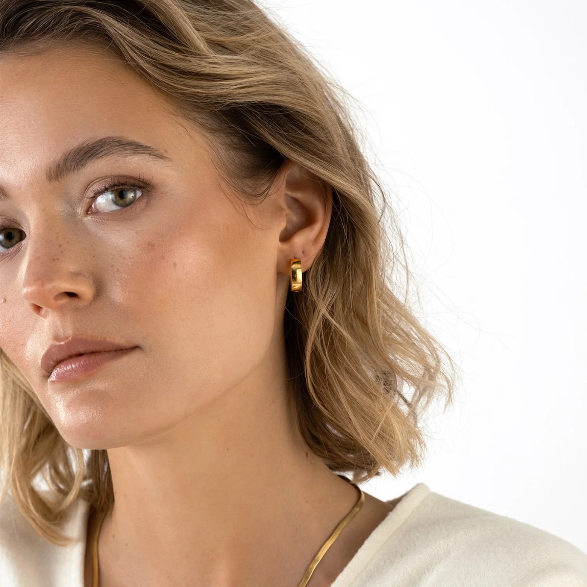 Et par små, glatte øreringer i gullfarget metall, vist mot hvit bakgrunn med svak refleksjon under smykkene.