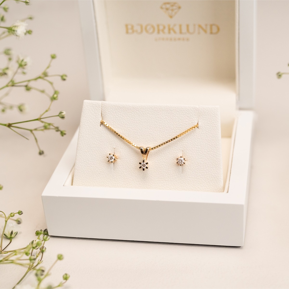 Et par små øredobber i gult metall med runde, klare steiner festet i seks klør, lagt på hvit bakgrunn.