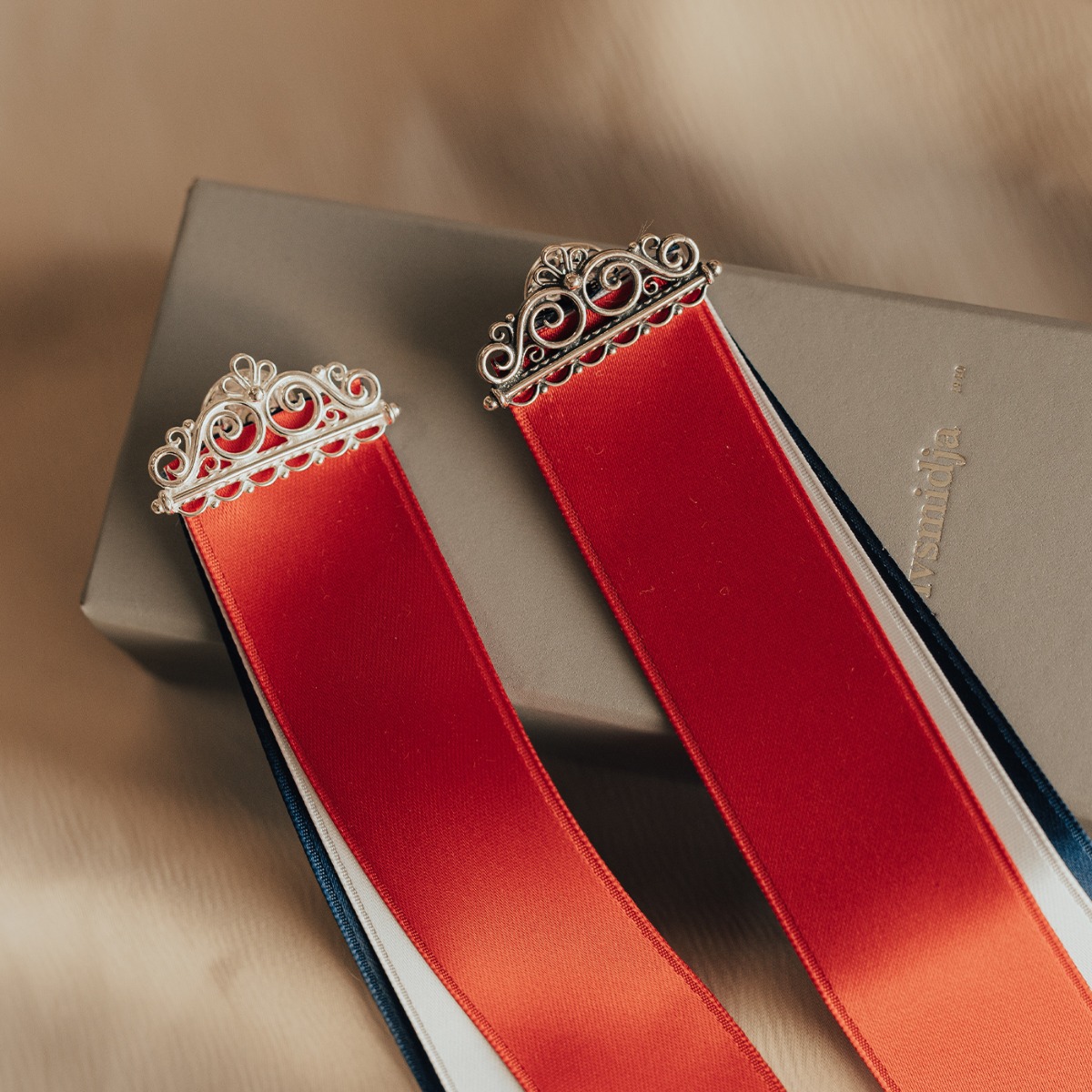 Rød, hvit og blå sløyfe med sølvfarget filigrandekor øverst, avbildet mot hvit bakgrunn.