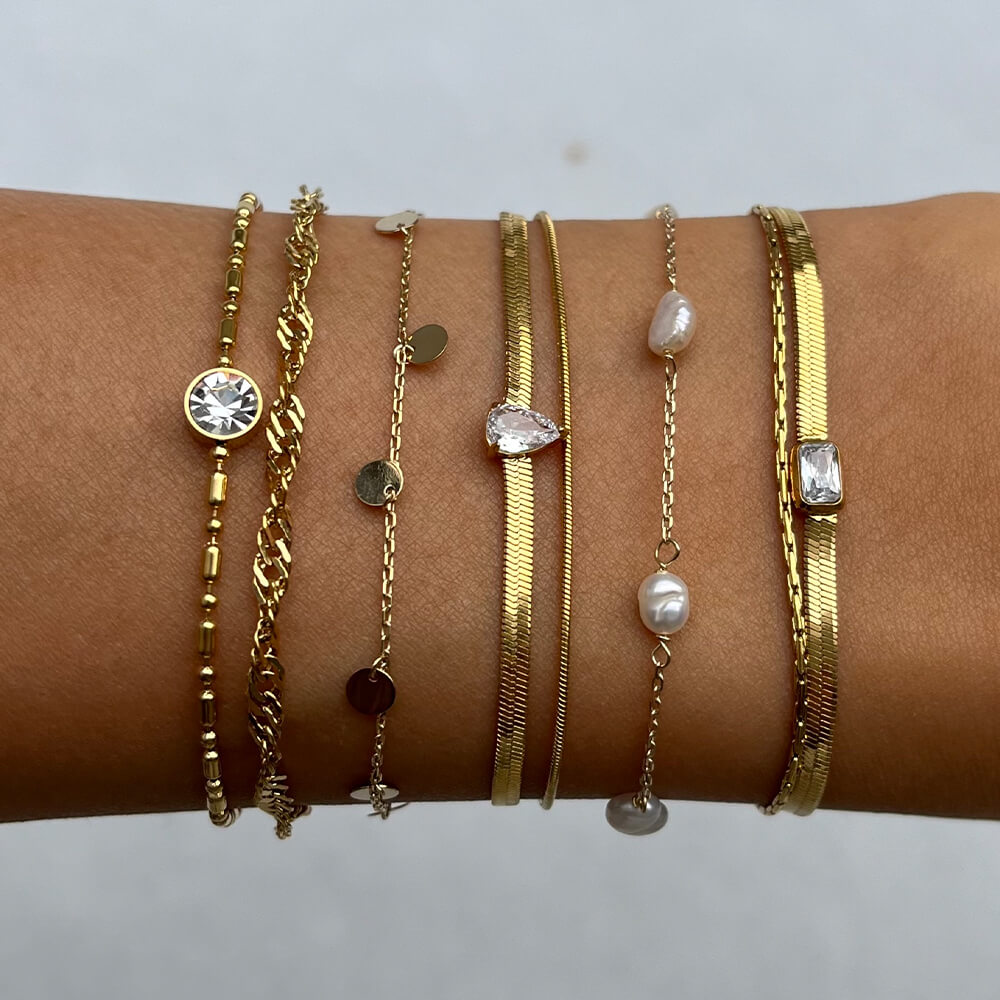 Armbånd i gullfarget lenke med to kjeder og en firkantet klar stein i midten, avbildet mot hvit bakgrunn.