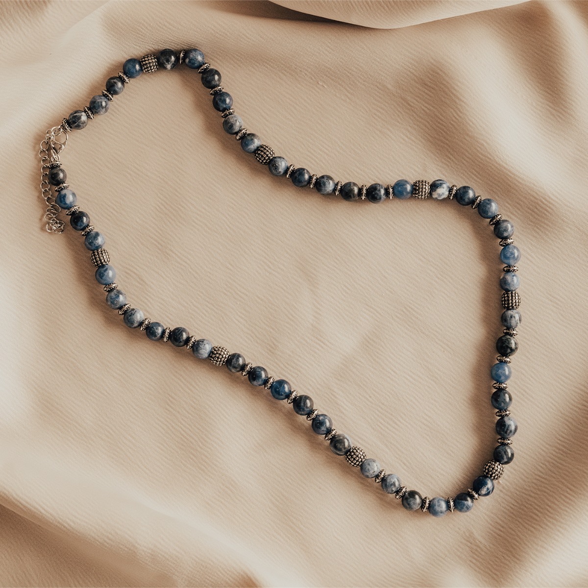 Halskjede med runde blå perler i ulike nyanser, dekorert med små sølvfargede ringer og enkelte større mønstrede sølvperler.