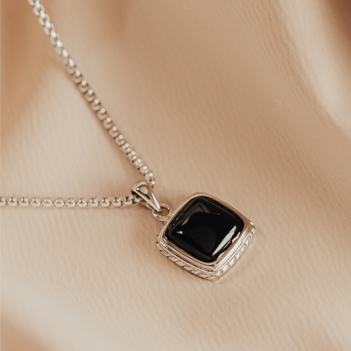 Sølvkjede med firkantet anheng som har en glatt, blank svart stein i midten, omgitt av dekorert sølvramme, mot hvit bakgrunn.