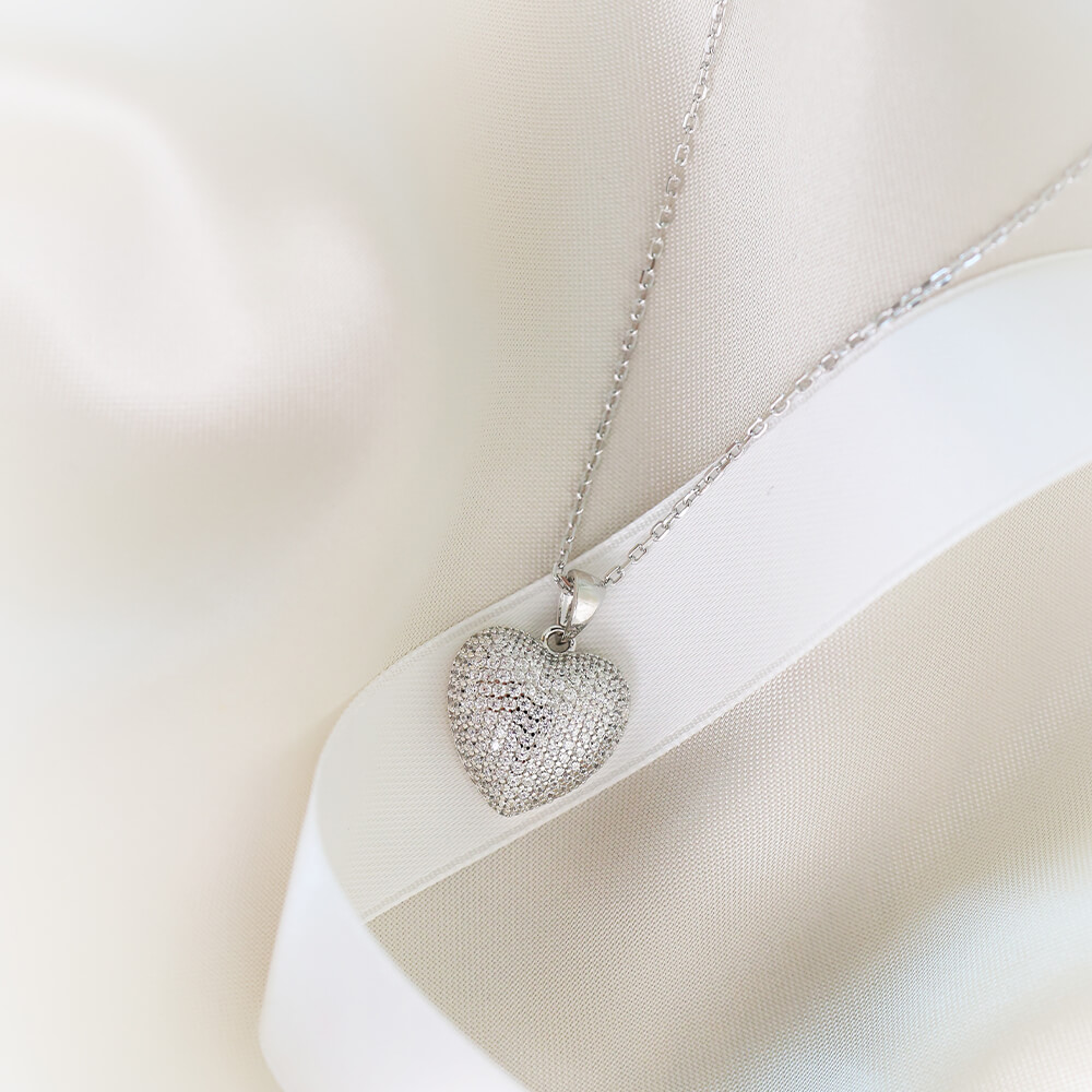 Sølvfarget halssmykke med halskjede og hjerteanheng dekket av små glitrende hvite stener.