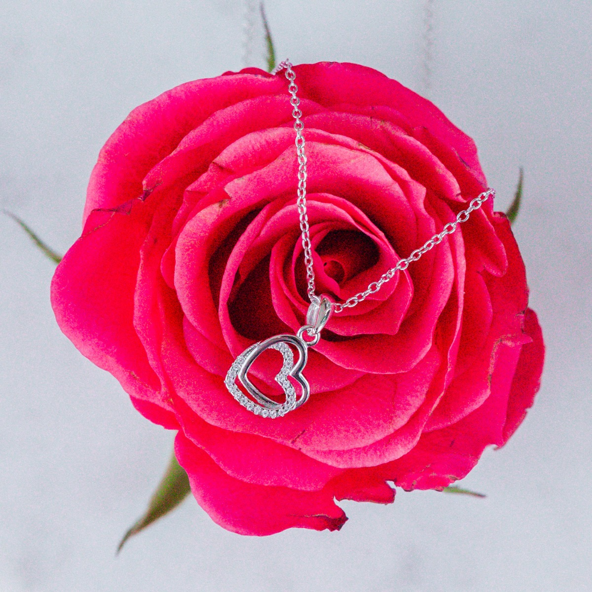 ølvfarget bokskjede-halssmykke med dobbelt hjerte-anheng der ett hjerte er dekorert med gjennomsiktige steiner mot hvit bakgrunn