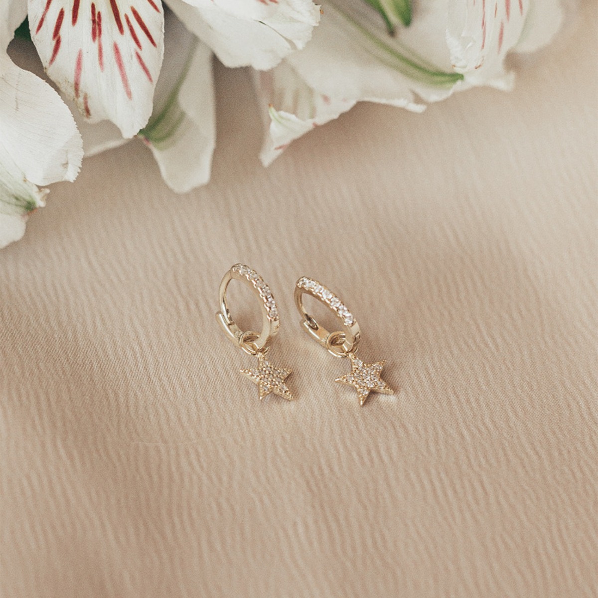 Små gullfargede hoops med hengende stjerneanheng, begge dekorert med klare glitrende steiner, avbildet mot hvit bakgrunn.