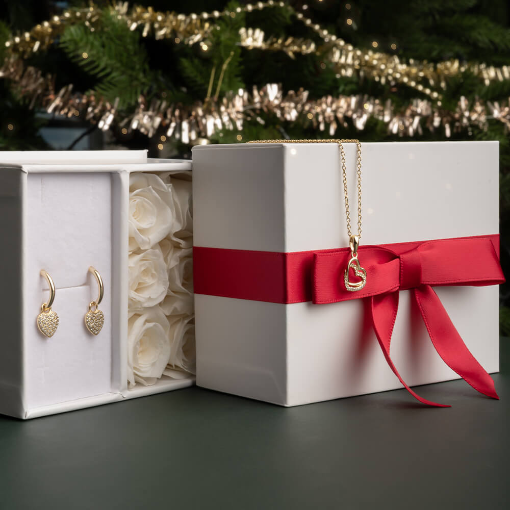 Par små gullbelagte creol-øredobber med hengende hjerte-anheng dekket av gjennomsiktige steiner vist mot hvit bakgrunn