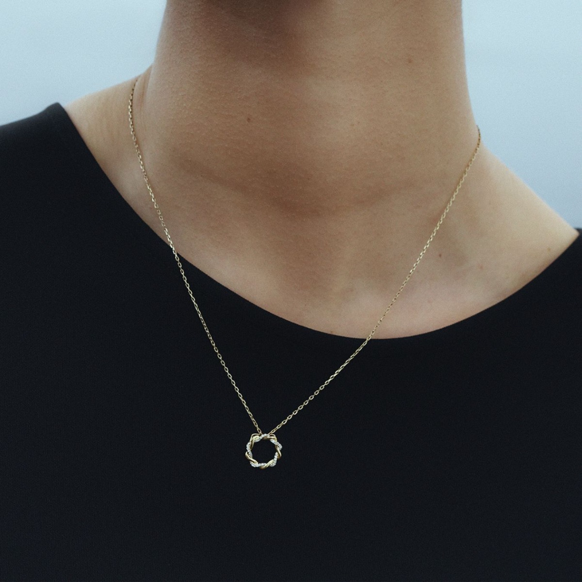 Gult gullkjede med rund vridd sirkel som anheng, vekselvis dekorert med små klare steiner, vist mot hvit bakgrunn.