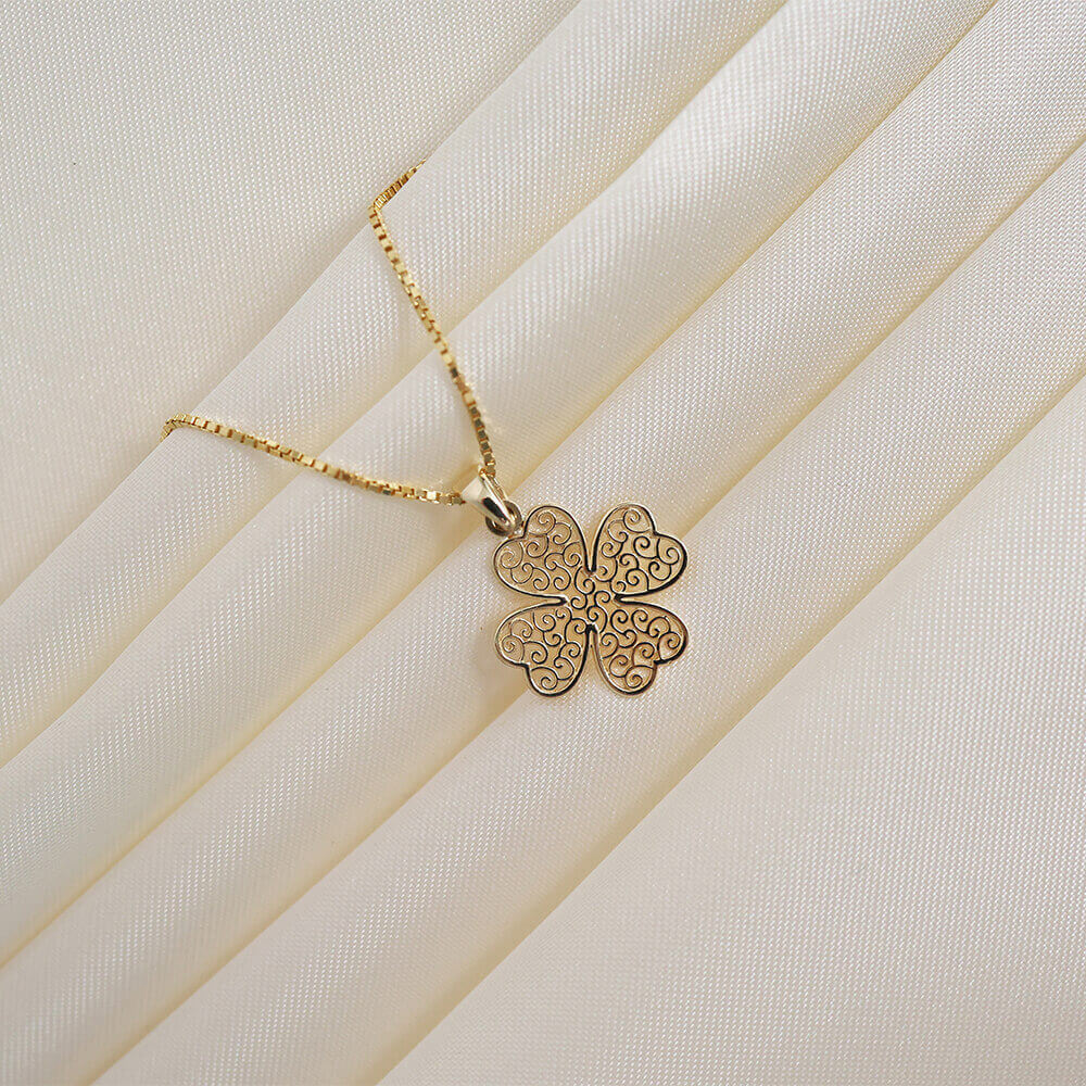 Gullfarget kjede med en firebladet anhengsformet medaljong med dekorativt mønster på hvit bakgrunn.