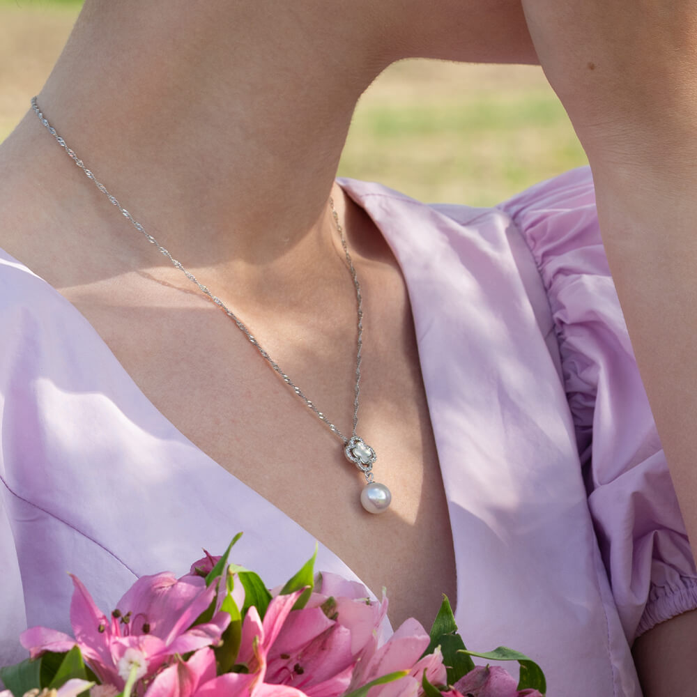 Et sølvkjede med et dekorativt kløver anheng med små steiner og en stor, rund perle hengende nederst, avbildet mot hvit bakgrunn.