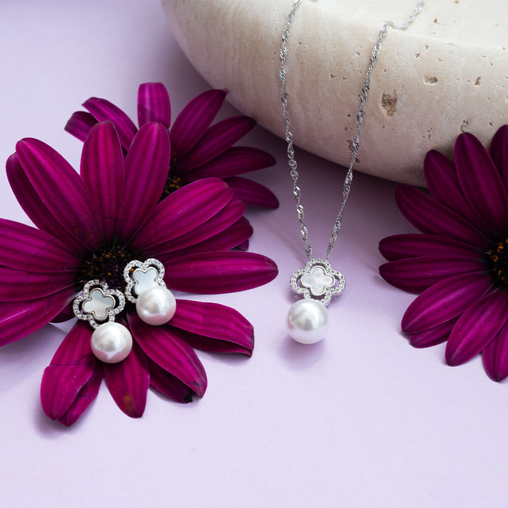 Et sølvkjede med et dekorativt kløver anheng med små steiner og en stor, rund perle hengende nederst, avbildet mot hvit bakgrunn.