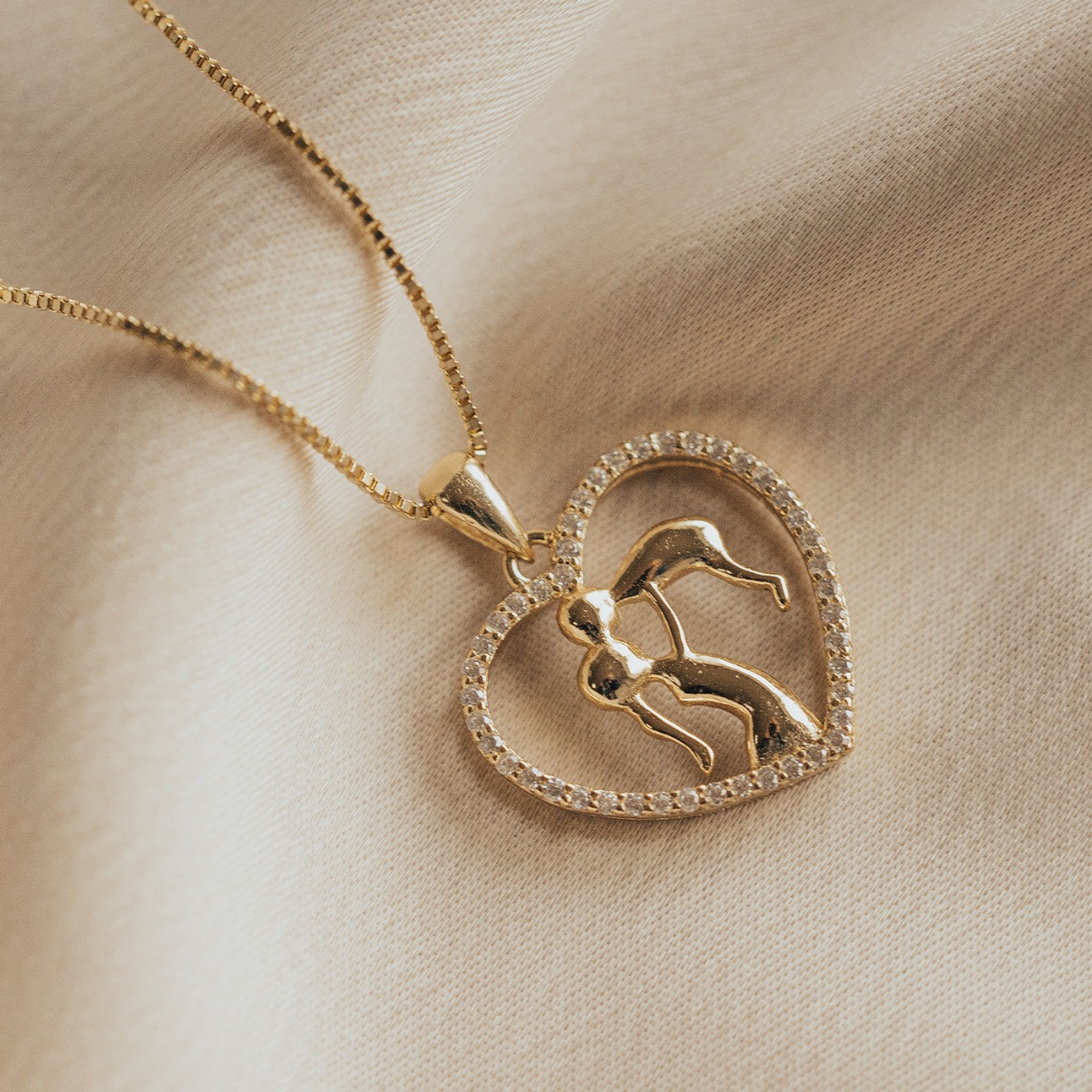 Gullfarget bokskjede med hjerte-anheng inneholdende mor og barn-motiv omkranset av gjennomsiktige steiner mot lys beige bakgrunn