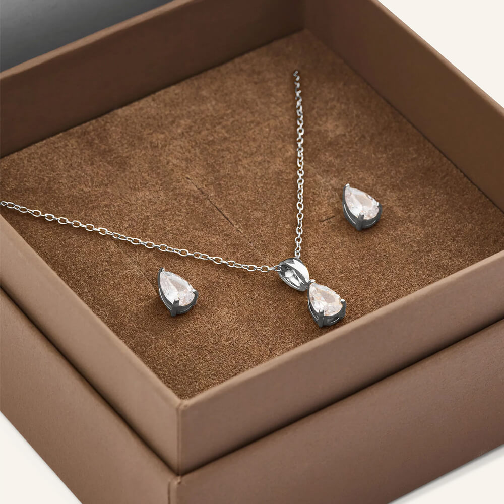 Sølvfarget smykkesett med kjede og to dråpeformede øredobber plassert i en brun eske med filtaktig innside.