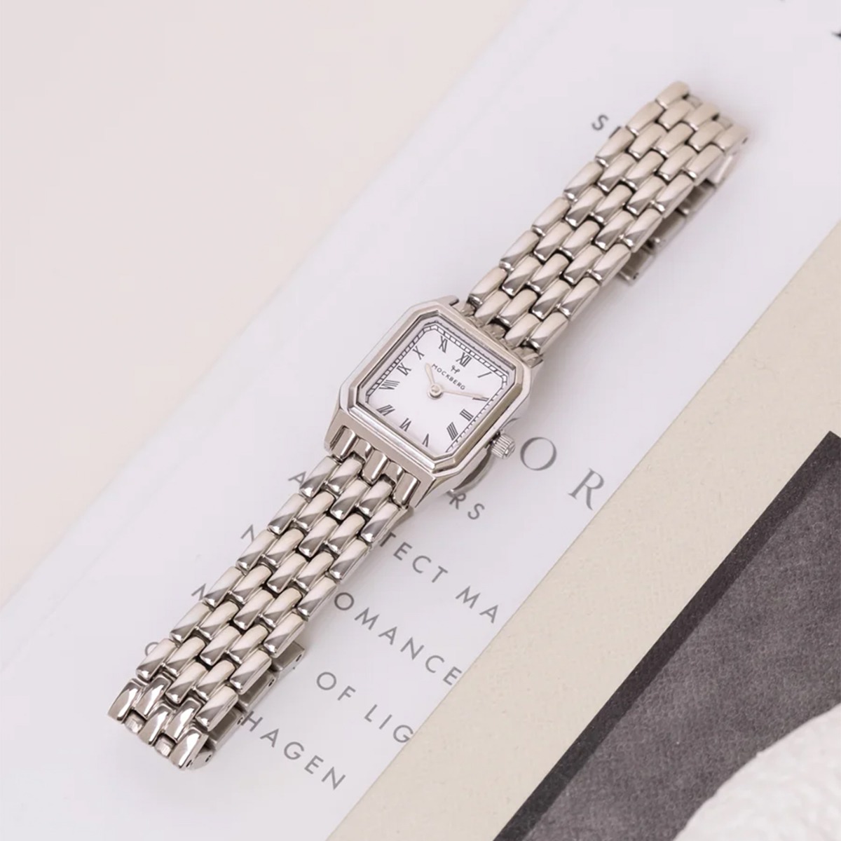 Sølvfarget Mockberg-klokke med hvit firkantet urskive og romertall på lenkearmbånd.