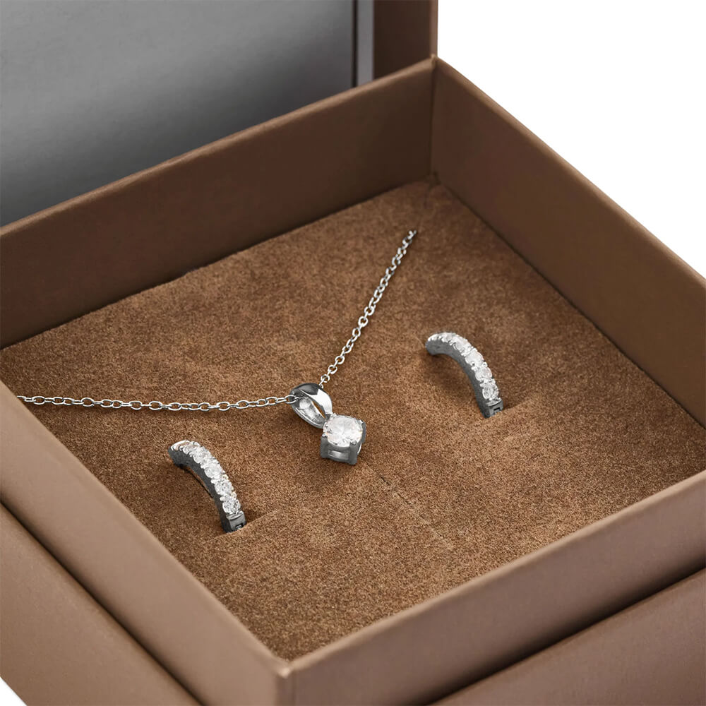 Smykkesett i sølv med halskjede og anheng med klar stein, samt matchende øreringer, presentert i brun gaveeske.