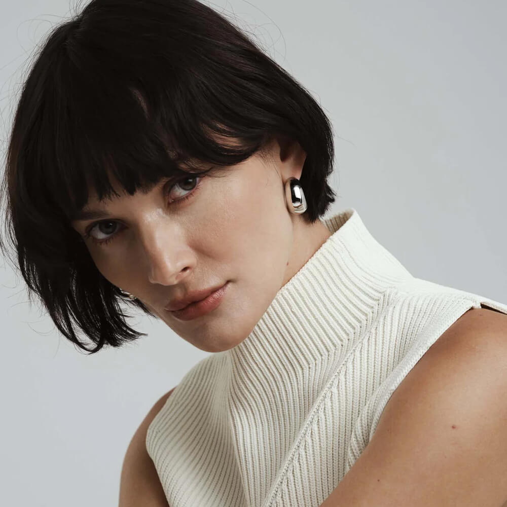 Et par små, runde øreringer i sølv med blank overflate, liggende side om side på hvit bakgrunn med lett refleksjon under.