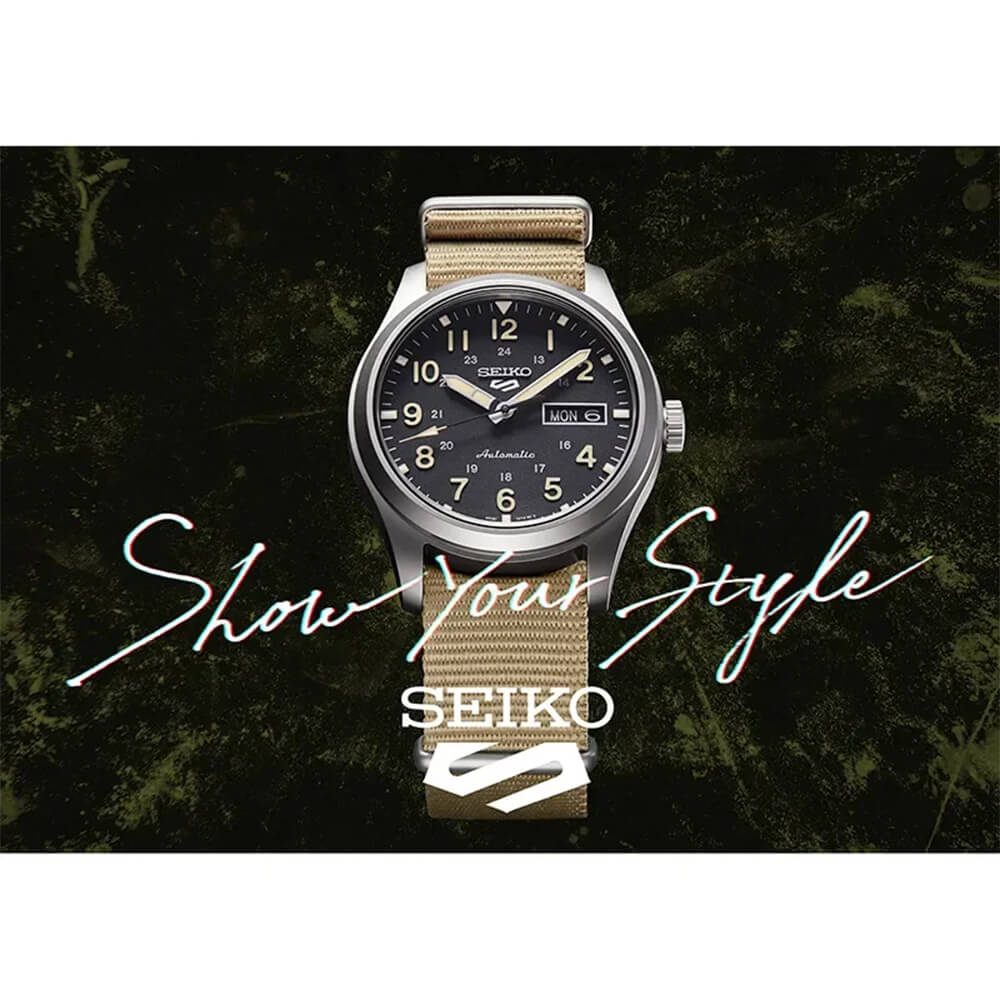 Armbåndsur med svart urskive, lyse tall, dag- og datovisning, sølvfarget urkasse og beige tekstilreim, sett forfra mot lys bakgrunn.