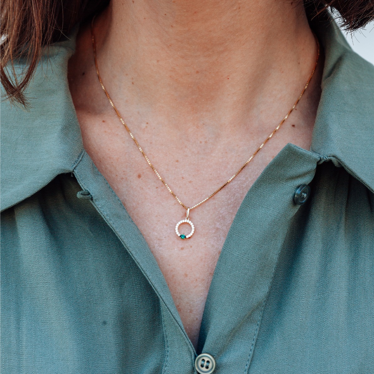 Gullfarget halskjede med lenkekjede og rundt anheng dekorert med små klare steiner og én grønn stein nederst, mot hvit bakgrunn.