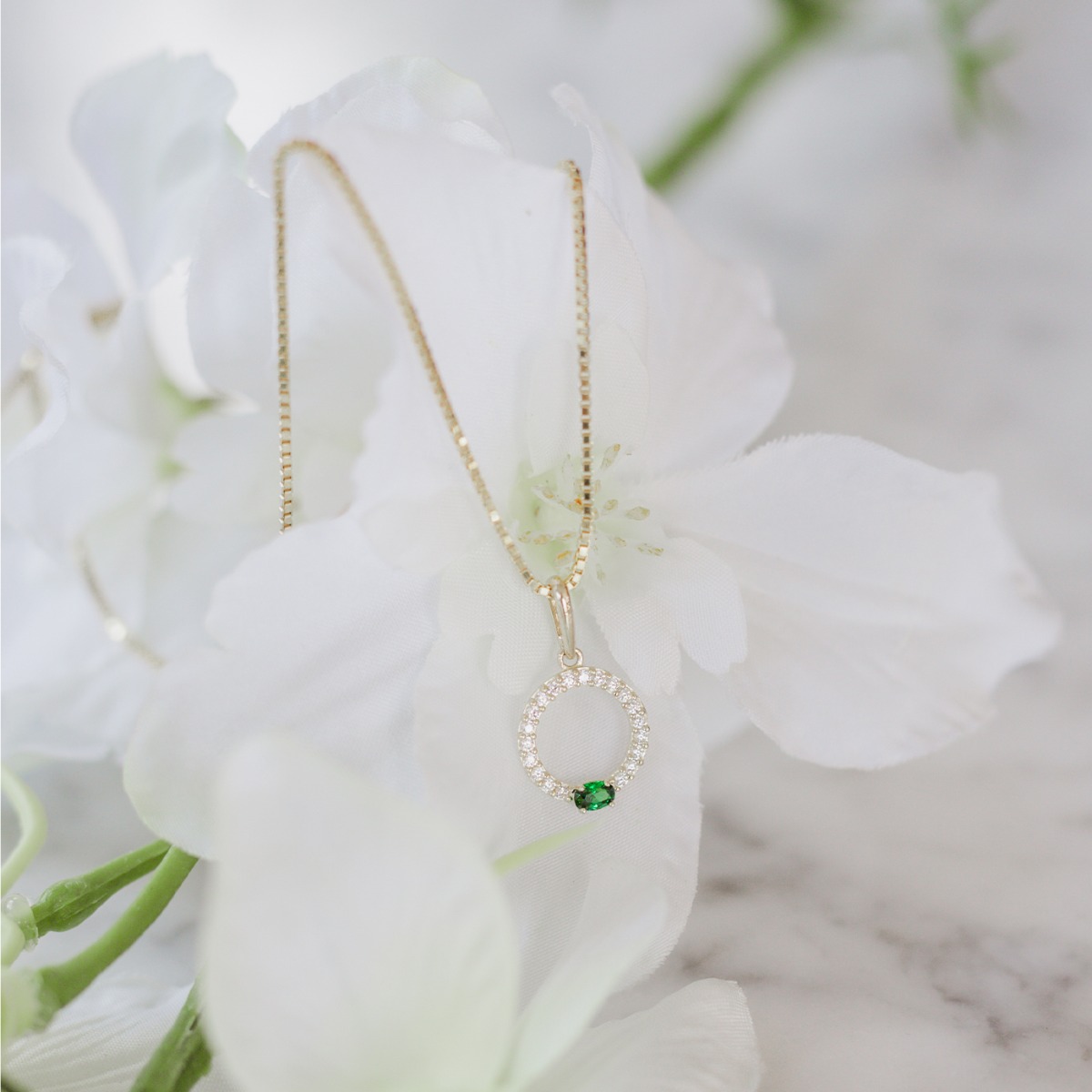 Gullfarget halskjede med lenkekjede og rundt anheng dekorert med små klare steiner og én grønn stein nederst, mot hvit bakgrunn.