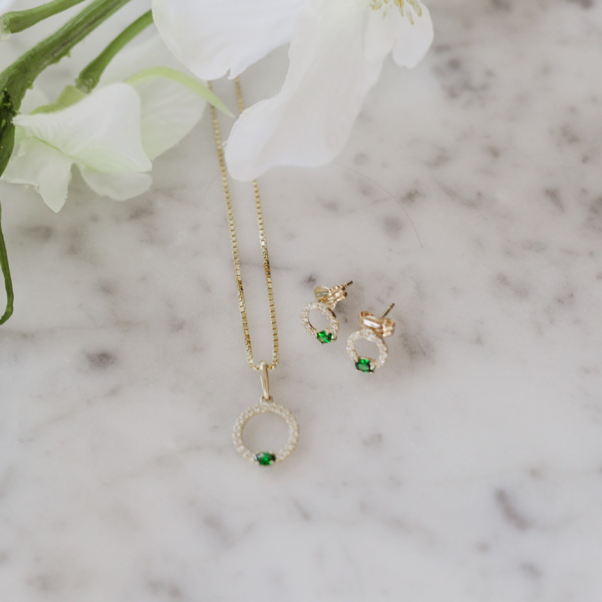 Par gullfargede ørestikker med oval form, små klare steiner rundt kanten og grønn stein nederst, fotografert mot hvit bakgrunn.